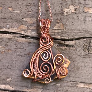 May include: A handcrafted pendant necklace featuring a copper-colored wire-wrapped design. The pendant has a dark gray and tan stone base with intricate wire swirls and a matching chain. The necklace is displayed against a weathered wooden surface.