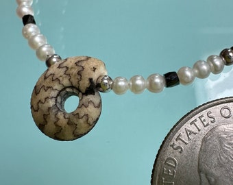 May include: A white pearl necklace with a black bead and a fossilized ammonite pendant. The necklace is next to a US quarter for size comparison.