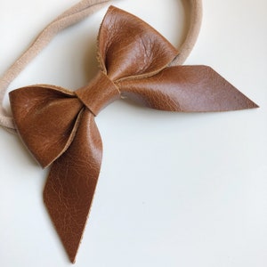 Puede incluir: Un lazo de cuero marrón atado a una diadema elástica de color nude.