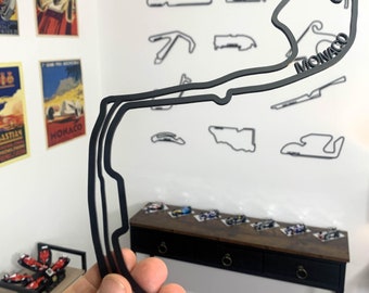 Puede incluir: Contorno negro del circuito de carreras de Mónaco con la palabra "MONACO" en la pista. Otros contornos de circuitos de carreras están en la pared blanca. Coches de carreras en miniatura sobre una mesa marrón oscuro.