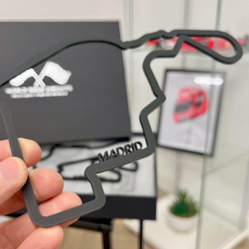 May include: A black, stylized outline of a race track, with the city name "MADRID" visible. The track is held in a hand. A black box with white text and a framed image of a red helmet are in the background.