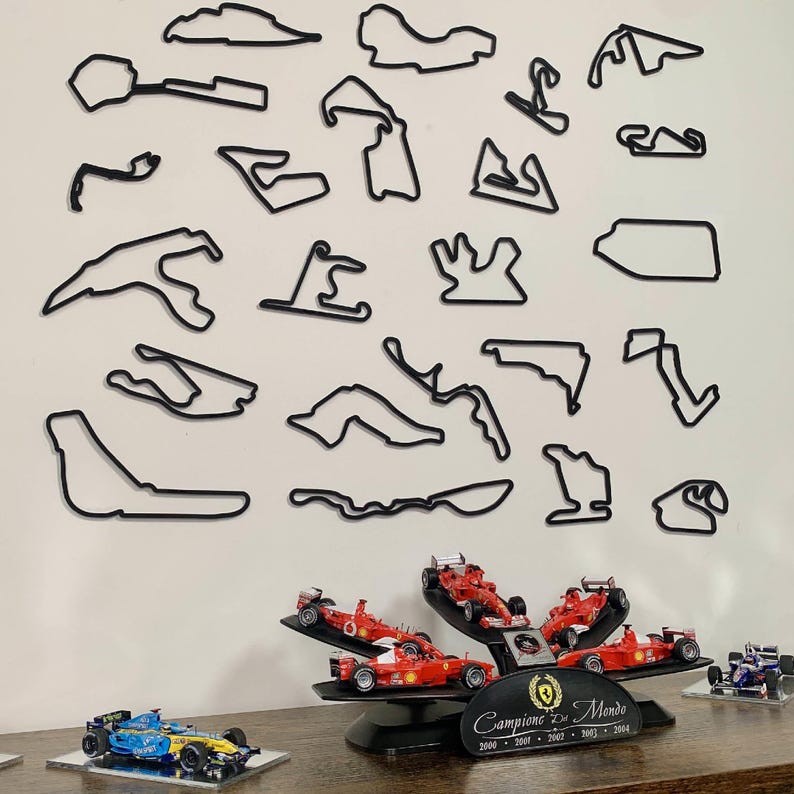 May include: A collection of black outline race track wall art and miniature Formula 1 race cars. The cars are red, blue, and yellow. A black display stand holds several red cars. The wall art is mounted on a white wall.