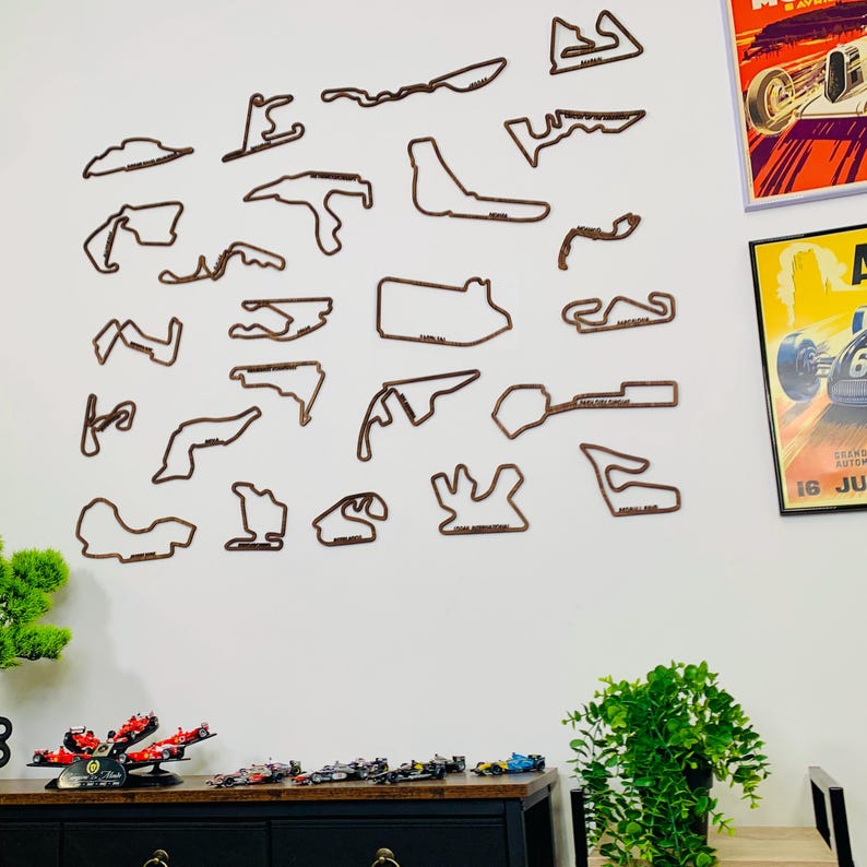May include: A collection of brown wooden race track outlines mounted on a white wall. The outlines vary in shape and size, with some labeled with track names. Decorative posters and a black cabinet with model race cars complete the scene.