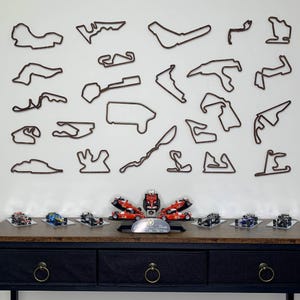 May include: A collection of miniature race cars in various colors, displayed on a dark wooden table. Above, brown outline wall art depicts race tracks. The cars are arranged with a central display piece and a silver plaque.