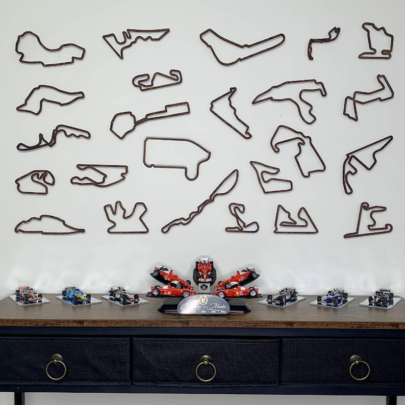 May include: A collection of miniature race cars in various colors, displayed on a dark wooden table. Above, brown outline wall art depicts race tracks. The cars are arranged with a central display piece and a silver plaque.
