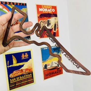 May include: A wooden cutout of the Circuit of the Americas racetrack, with the text "CIRCUIT OF THE AMERICAS" carved into the wood. The cutout is held in front of vintage racing posters.