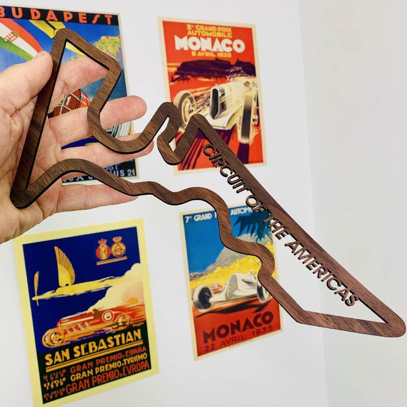 May include: A wooden cutout of the Circuit of the Americas racetrack, with the text "CIRCUIT OF THE AMERICAS" carved into the wood. The cutout is held in front of vintage racing posters.