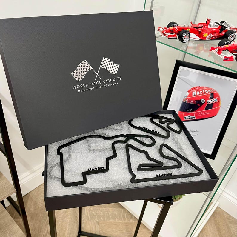 May include: A black box set featuring black race track outlines, including Madrid and Bahrain, with the text "WORLD RACE CIRCUITS Motorsport Inspired Artwork." A framed picture of a red racing helmet and a red race car are also visible.