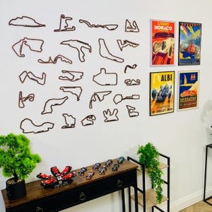 Può includere: Un'esposizione a parete con contorni di circuiti di gara in legno marrone e manifesti di corse vintage. Un tavolino nero ospita modellini di auto da corsa e un fiore decorativo. I manifesti includono le parole "Monaco", "Albi" e "San Sebastian".