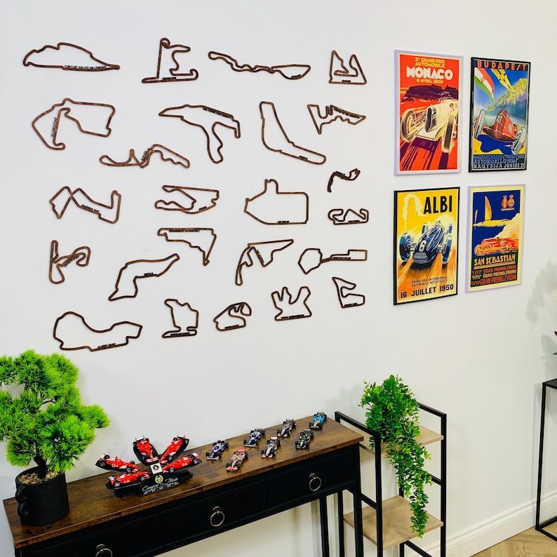 May include: A wall display featuring brown wooden race track outlines and vintage racing posters. A black console table holds miniature race cars and a decorative flower. The posters include the words "Monaco", "Albi", and "San Sebastian".