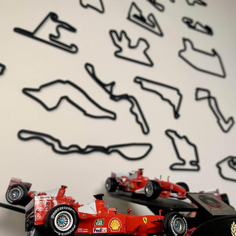 May include: A display featuring red and white Formula 1 race cars with sponsor logos, positioned in front of black outline track layouts on a white wall. The cars have detailed features, including tires and aerodynamic elements.