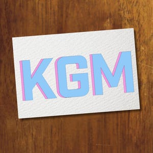 May include: A white card with the letters "KGM" in light blue, outlined in pink, against a dark wood grain background. The letters appear to have a 3D effect, creating a shadow. The card is slightly angled.