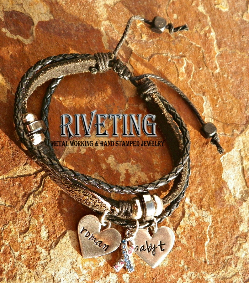 Peut inclure: Un bracelet en cuir noir avec un charme argent&eacute; qui dit "baby" et un charme en forme de c&oelig;ur argent&eacute; qui dit "roman". Le bracelet a &eacute;galement une perle argent&eacute;e et un charme en forme de plume argent&eacute;. Le bracelet est sur un fond brun.
