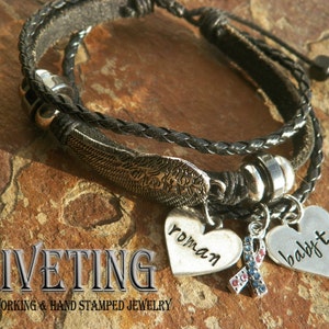 Peut inclure: Un bracelet en cuir noir &agrave; trois brins avec des breloques argent&eacute;es. Les breloques comprennent un c&oelig;ur avec le mot "roman" dessus, un c&oelig;ur avec le mot "baby" dessus et un breloque avec un ruban bleu et rouge. Le bracelet est sur une surface textur&eacute;e brune.