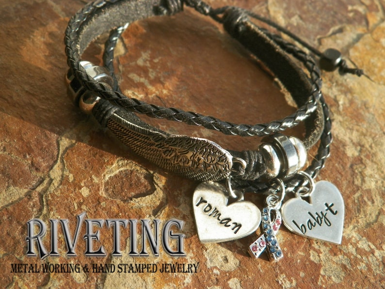 Peut inclure: Un bracelet en cuir noir &agrave; trois brins avec des breloques argent&eacute;es. Les breloques comprennent un c&oelig;ur avec le mot "roman" dessus, un c&oelig;ur avec le mot "baby" dessus et un breloque avec un ruban bleu et rouge. Le bracelet est sur une surface textur&eacute;e brune.