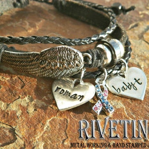 Peut inclure: Un bracelet en cuir noir avec un charme d'aile argent&eacute;, un charme de c&oelig;ur avec le mot "roman" dessus, un charme de c&oelig;ur avec le mot "baby" dessus et un charme avec un ruban bleu et rouge. Le bracelet est sur une surface de roche brune. Le texte "RIVETING METAL WORKING & HAND STAMPED JEWELRY" est sous le bracelet.
