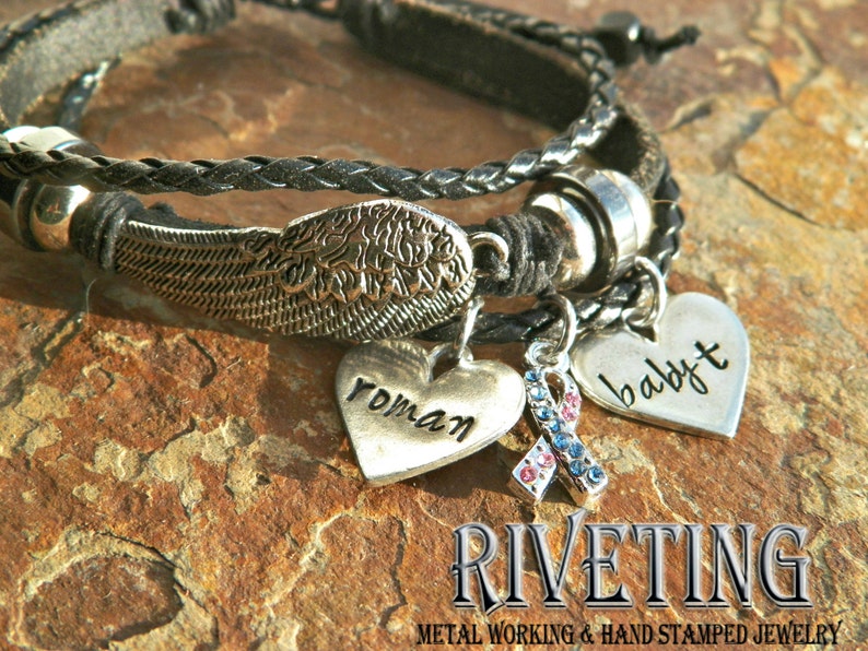 Peut inclure: Un bracelet en cuir noir avec un charme d'aile argent&eacute;, un charme de c&oelig;ur avec le mot "roman" dessus, un charme de c&oelig;ur avec le mot "baby" dessus et un charme avec un ruban bleu et rouge. Le bracelet est sur une surface de roche brune. Le texte "RIVETING METAL WORKING & HAND STAMPED JEWELRY" est sous le bracelet.