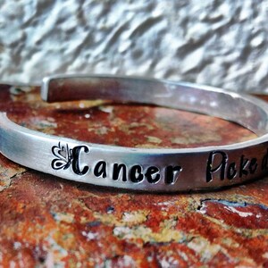 May include: A silver cuff bracelet engraved with the words "Cancer Picked" in black. A butterfly design precedes the word "Cancer". The bracelet is shown on a textured, reddish-brown surface. The bracelet is a piece of jewellery.