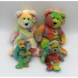 May include: Two large and two small plush teddy bears with tie-dye patterns in pastel and vibrant colors. Each bear features a black plastic nose, black eyes, and a circular patch on the chest displaying a multi-colored peace sign symbol.