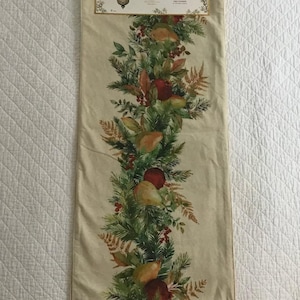 May include: A cream-coloured rectangular table runner with a central garland design of green pine needles, red apples, and pears. The runner is suspended from a silver hanger.