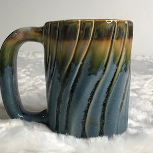 Puede incluir: Una taza de cerámica con un diseño texturizado único. La taza presenta un asa y un cuerpo con ranuras verticales en forma de onda. El color pasa de un azul oscuro en la base a verde y dorado en la parte superior, creando un efecto degradado.