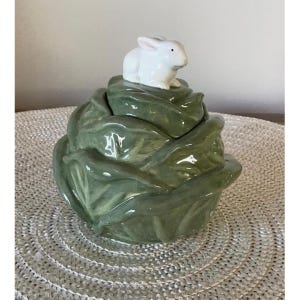 May include: A ceramic lidded jar shaped like a head of green cabbage with a white rabbit figurine perched on top of the lid. The cabbage leaves feature realistic molded textures and a glossy finish, while the rabbit has smooth white surfaces.