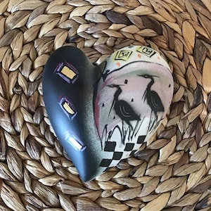 May include: Heart-shaped ceramic ornament. One side is black with gold rectangles, the other features two birds in a pink and white landscape with a black and white checkered pattern. Set against a woven background.