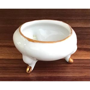 May include: A small, white ceramic bowl with a rounded, bulbous shape. The rim is accented with a thin gold-colored trim, and the bowl is supported by three short, curved legs that feature gold-colored tips at the base of each foot.