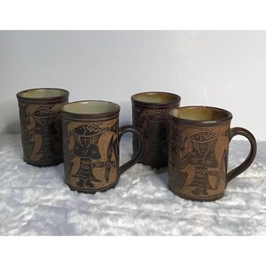 May include: Four brown ceramic mugs with handles. Each mug features a carved design of a person with a hat and fish. The mugs have a rustic, handcrafted appearance, with variations in colour and texture. The mugs are set against a white background.
