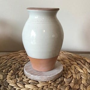 Puede incluir: Un jarrón de cerámica blanca con base y borde de terracota. El jarrón se asienta sobre un pequeño soporte de madera redondo y blanqueado. El jarrón tiene un acabado liso y brillante y una forma clásica y redondeada. El fondo es de color neutro.