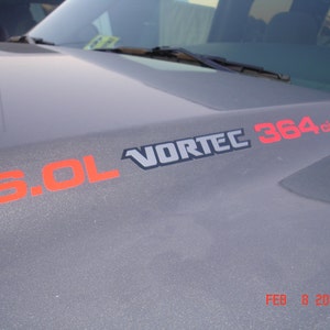May include: Close-up of a gray vehicle hood with the text "VORTEC" in silver and black, and "6.0L 364ci" in orange and red. The date "FEB 8 2007" is visible in the bottom right corner.