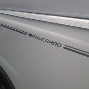 Op de afbeelding: Close-up van een witte truckbed met zwarte accentstrepen en het woord "SILVERADO" in zwarte letters. Het Chevrolet-logo is ook zichtbaar.