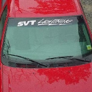 May include: A red car with a windshield banner that reads "SVT LIGHTNING" in white lettering. The car has a glossy finish and a windshield with wipers. The side mirror is visible on the left side of the car.