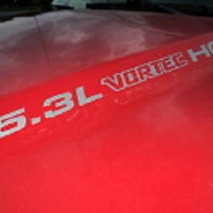 May include: Bright red vehicle hood with the text "5.3L VORTEC HO" in silver lettering. The hood appears to be slightly curved, and the surface reflects light. The image focuses on the engine specifications.