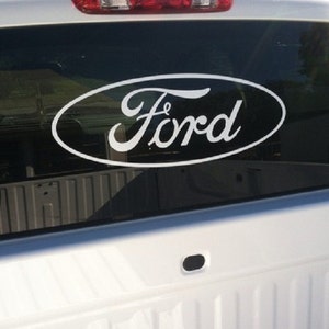 May include: White Ford logo sticker on a vehicle's rear window. The Ford logo is in a white oval shape with the word "Ford" in a cursive font. The vehicle's white tailgate and rear window are visible.
