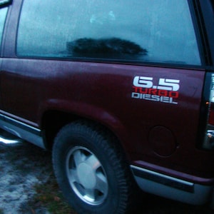 Puede incluir: Un SUV granate con un estribo cromado y un neumático negro. El vehículo tiene un emblema "6.5 Turbo Diesel" en blanco y rojo. La ventana está ligeramente tintada y el vehículo parece estar al aire libre.