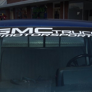 May include: A blue truck with a white "GMC TRUCK MOTORSPORTS" decal across the top of the rear window. The decal has a checkered flag design on the left side. The interior of the truck is visible.