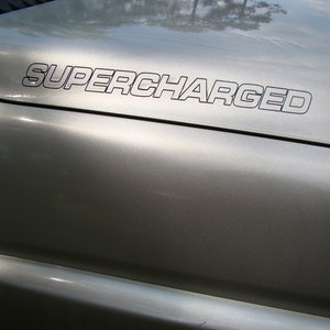 Puede incluir: Primer plano del capó plateado de un vehículo con la palabra "SUPERCHARGED" en letras negras delineadas. La superficie metálica brillante refleja el entorno. El texto está centrado y situado en la parte superior del capó.