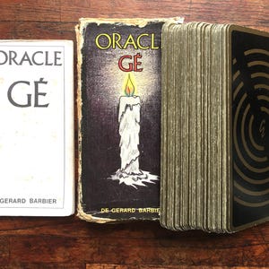 Puede incluir: Un folleto de instrucciones blanco con la etiqueta ORACLE GÉ GERARD BARBIER, una caja negra con una ilustración de una vela encendida y la etiqueta ORACLE GÉ DE GERARD BARBIER, y una baraja con dorso negro, patrón circular dorado y la letra G.