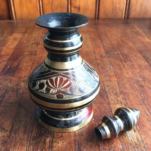 Pode incluir: Um pequeno recipiente de metal bojudo com borda alargada, apresentando um acabamento preto com faixas horizontais douradas e padrões florais gravados em vermelho e ouro. Uma tampa cónica a condizer com faixas semelhantes encontra-se ao lado.