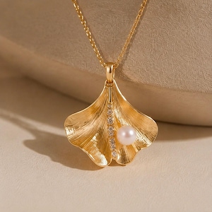 May include: A gold-toned necklace featuring a ginkgo leaf-shaped pendant with a textured surface. A vertical row of small, clear, sparkling stones runs down the center of the leaf, accompanied by a single, smooth, round white pearl.