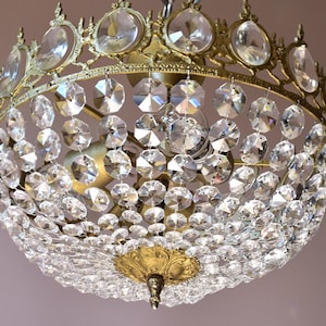 Peut inclure: Un lustre vintage avec une monture en métal doré et une multitude de pendentifs en cristal clair et facetté. Les cristaux sont disposés en forme sphérique, reflétant la lumière. Le luminaire a un design orné et décoratif.