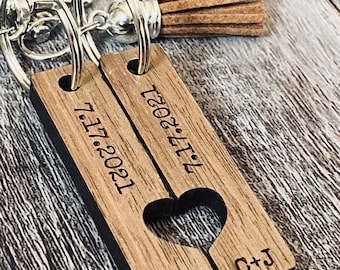 May include: A wooden keychain with two pieces that fit together to form a heart. The pieces are engraved with the date "7.17.2021" and "7.17.2021" and the initials "J+C" and "C+J". The keychain has a brown tassel.