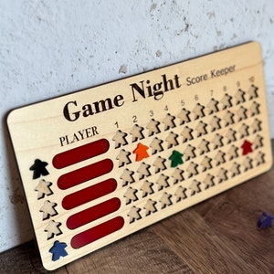 May include: Wooden game night score keeper with the words "Game Night Score Keeper" printed on the top. The score keeper has spaces for four players, with red slots and wooden player pieces. Colored player pieces include black, blue, orange, green, and red.