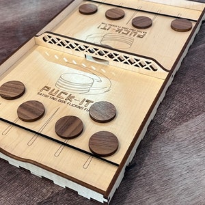 May include: A wooden game called "Puck-It!" with the text "Satisfying Disk Flicking Fun" printed on the surface. The game features a wooden frame with a series of wooden pegs and a set of wooden pucks.