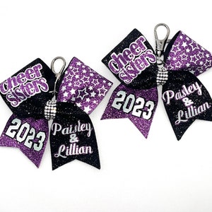 May include: Two black and purple cheer bows with silver glitter and white stars. The bows have the text "cheer sisters" and "2023" on them. The bows also have the names "Paisley & Lillian" on them.