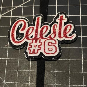 May include: A black headband with a glittery, custom-made name tag. The name "Celeste" is written in red glitter with a white outline, and the number "6" is below it. The name tag has a black border.
