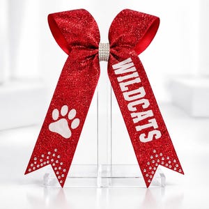 May include: A red glitter cheer bow featuring a central rhinestone band. The left tail displays a white paw print graphic, while the right tail features the white text WILDCATS. Both tails are accented with small clear rhinestones at the bottom.