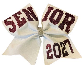May include: White glitter cheerleading bow with maroon glitter text that reads "SENIOR 2021". The bow is tied in the centre with a silver rhinestone band. A festive accessory for a senior.