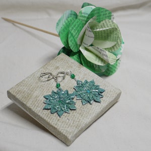 May include: A pair of earrings with green paper flower designs and silver hooks. The earrings are displayed on a small canvas with a printed pattern.